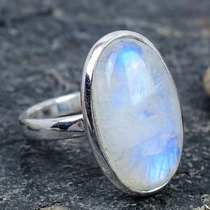 May include: A silver ring with a large, oval moonstone. The moonstone displays a milky white base with iridescent blue hues. The ring's band is polished silver.
