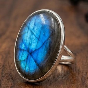 May include: A silver ring featuring a large, oval-shaped labradorite gemstone. The stone displays a prominent iridescent blue flash across its surface, set within a smooth, polished silver bezel. The band is split into two thin, metallic silver arms.