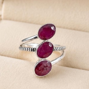 Könnte beinhalten: Ein silberner Ring mit drei ovalen, tiefroten Edelsteinen. Das Band des Rings teilt sich in zwei Teile, wobei sich eine Seite um die Edelsteine wickelt und die andere das Band bildet. Der Ring wird in einer cremefarbenen Schmuckschatulle präsentiert.