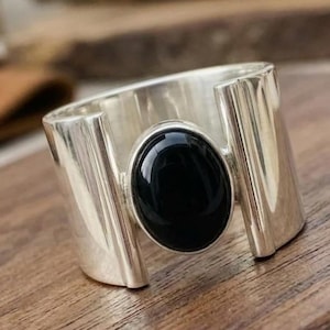 May include: A wide silver ring featuring a polished, oval-shaped black stone set in the centre. The band has a smooth, metallic finish with a distinct open-gap design on both sides of the central stone, creating a bold and structured appearance.