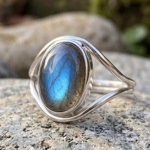 Op de afbeelding: Een zilveren ring met een ovale, iriserende blauwe edelsteen. De band van de ring heeft een gesplitst ontwerp en de edelsteen is gezet in een bezel setting. De ring wordt getoond op een grijze rots.