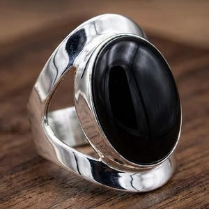 May include: A polished silver ring featuring a large, smooth, oval-shaped black stone set in the center. The band has a wide, open, and fluid design with a reflective metallic surface that curves around the central black gemstone setting.