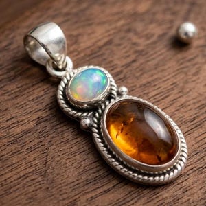 May include: A silver pendant featuring two gemstones: an oval amber-colored stone and a smaller, oval opal with iridescent hues. The pendant has a detailed, rope-like border and a bail for a chain. The pendant is displayed on a wooden surface.