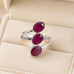 May include: A silver bypass ring featuring three oval-cut, deep pinkish-red rubies arranged in a vertical row. The metal band has a textured, ribbed finish along the sides, and the ring is displayed inside a cream-colored velvet jewelry box.