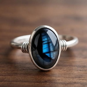Può includere: Un anello in argento con una pietra di labradorite di forma ovale, caratterizzata da una base grigio scuro e un vivido riflesso blu. La fascia è realizzata in filo metallico liscio che si avvolge attorno alla base della montatura su entrambi i lati.