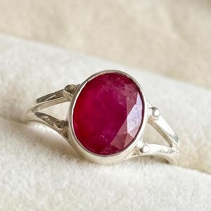 Könnte beinhalten: Ein silberner Ring mit einem großen, oval geschliffenen Rubin. Der Rubin hat eine tiefe, lebendige rote Farbe und ist in einem schlichten, eleganten Silberband gefasst. Der Ring wird in einer weißen Schmuckschatulle präsentiert.