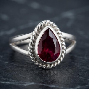 May include: A silver ring featuring a pear-shaped, deep red faceted gemstone set in a bezel. The stone is surrounded by a twisted rope-style metal border. The band of the ring is split into two separate silver strands that meet at the sides.