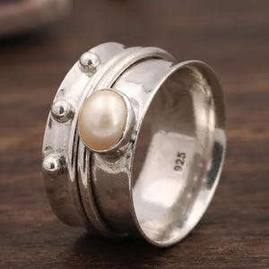 May include: A wide sterling silver spinner ring featuring a central oval white pearl set in a bezel. The band has a hammered texture with three small silver spheres on one side and is stamped with the number 925 on the inner surface.