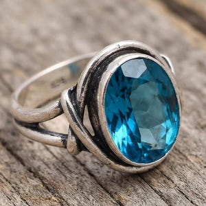 May include: A silver ring featuring a large, oval-cut, faceted blue gemstone set in a polished metal band. The setting includes a decorative, twisted metal wire design that wraps around the base of the vibrant blue stone, creating a textured look.