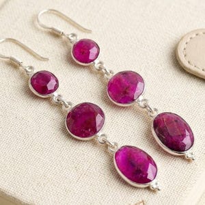 May include: Dangle earrings with magenta gemstones set in silver. The earrings feature oval and round gems, connected by silver links. The faceted gemstones create a sparkling effect. The earrings' design is a cascade of gems, adding a touch of elegance.