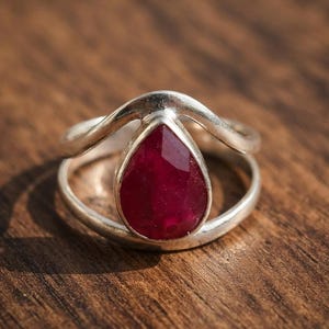 May include: A silver ring featuring a teardrop-shaped, faceted deep red gemstone set in a split-shank band. The metal band curves upwards to frame the stone, creating a layered appearance with a polished finish and a vibrant, dark red centre.