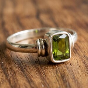 May include: A silver ring featuring a rectangular, olive-green gemstone. The ring has a simple band with decorative elements on the sides of the gemstone setting. The gemstone is approximately 1.3 cm in length.