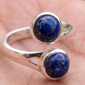 May include: A silver ring with two round, deep blue lapis lazuli stones. The ring's band curves around the finger, with one stone at the top and the other at the bottom. The stones have flecks of gold.