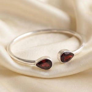 Puede incluir: Una pulsera abierta de plata con dos piedras preciosas de granate rojo oscuro en forma de lágrima en cada extremo. La banda de metal pulido tiene un perfil liso y redondeado, y las dos piedras están engastadas en sencillos biseles de plata.