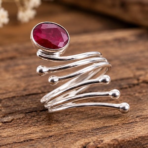 Puede incluir: Un anillo de plata con una piedra preciosa de color rojo intenso, forma ovalada y tallado facetado en la parte superior. La banda tiene un diseño de espiral multicapa que se envuelve alrededor del dedo, con cada extremo del hilo de plata acabado en una pequeña bola pulida.