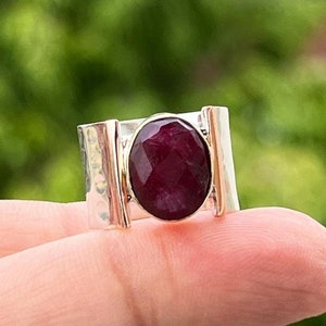 May include: A silver ring featuring a large, oval-shaped, dark red gemstone set in the centre. The ring band is textured and has two vertical bars on either side of the stone.