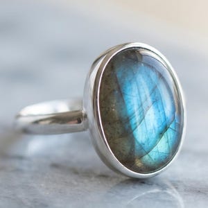 May include: A silver ring featuring a smooth, oval-shaped labradorite gemstone. The stone displays a prominent iridescent blue flash across its grey surface, set within a simple, polished metal bezel band that encircles the finger.