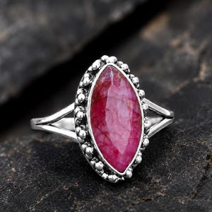 Op de afbeelding: Een zilveren ring met een gefacetteerde, donkerroze edelsteen in marquise-slijpvorm, gezet in een decoratieve zetting met kleine metalen bolletjes. De ringband splitst zich in twee dunne, gepolijste zilveren armen die de zetting verbinden.