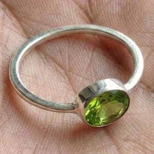 May include: A silver ring with a bright green oval peridot gemstone. The stone is set in a polished silver band. The ring is displayed on a hand, showcasing its design. The peridot is a vivid, light green colour.