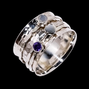 May include: A silver spinner ring with a hammered texture. The ring features a small, round, light blue gemstone, a small heart-shaped silver accent, and a small, round, purple gemstone. The ring is set against a black background.