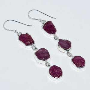 Peut inclure: Boucles d'oreilles pendantes composées de trois pierres de rubis brutes, rouge foncé, serties dans du métal argenté. Les boucles d'oreilles ont une fermeture à crochet simple, mettant en valeur la beauté naturelle des pierres.