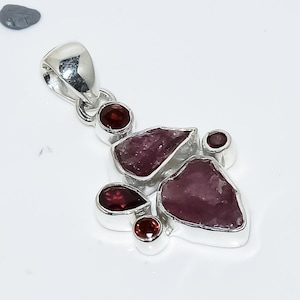 May include: A silver pendant featuring a cluster of irregularly shaped, deep pink gemstones and smaller, round, dark red gemstones. The pendant has a silver bail for hanging and is set against a white background.