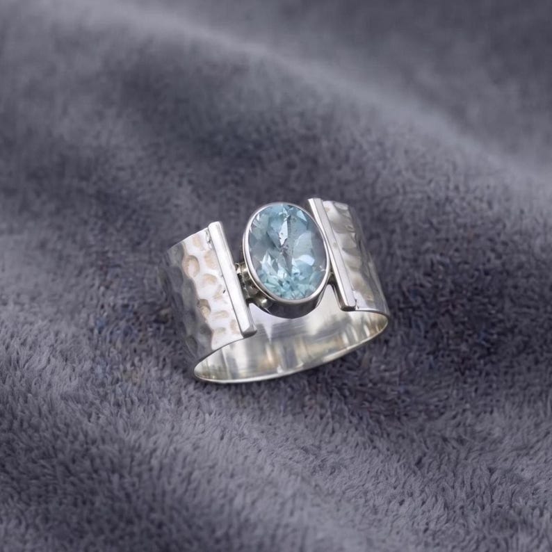 May include: A wide silver band ring featuring a central oval-shaped light blue gemstone. The metal surface of the ring displays a distinct hammered texture on the sides, providing a contrast to the smooth, polished finish of the setting.