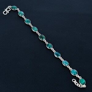 May include: A silver bracelet with oval, vivid green gemstones. The gemstones are connected by a delicate silver chain and clasp. The bracelet is shown against a dark background, accentuating the jewellery's sparkle and design.
