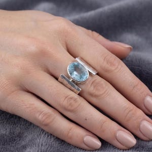 May include: A sterling silver ring featuring a large, oval-cut light blue gemstone set in the center. The band consists of two wide, flat, hammered silver bars that extend from the central stone setting, creating a bold and open geometric design.