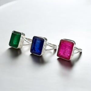 Może przedstawiać: Trzy srebrne pierścionki z różnokolorowymi kamieniami szlachetnymi. Pierścionki mają prosty design z kwadratowym kamieniem szlachetnym w środku. Kamienie szlachetne są zielone, niebieskie i różowe.