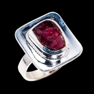 May include: A silver ring with a large, irregularly shaped, deep red gemstone set in a square frame. The ring has a polished surface and a simple band. The gemstone's colour contrasts with the silver setting.