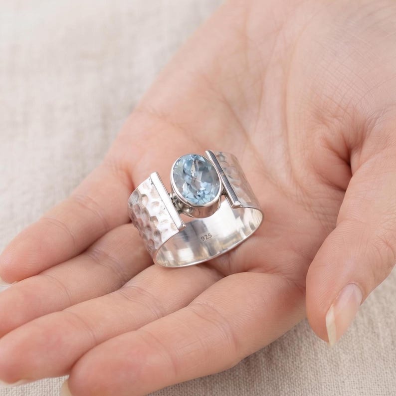 May include: A wide sterling silver ring featuring a hammered texture on the band and a central oval-cut light blue gemstone. The interior of the band is clearly stamped with the hallmark 925, indicating the silver content of the jewelry piece.