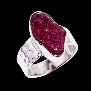 Könnte beinhalten: Ein Silberring mit einem großen, unregelmäßig geformten, tiefroten Edelstein. Der Edelstein ist in einer glatten Lünette gefasst, und das Band hat ein strukturiertes, gehämmertes Aussehen. Der Ring ist vor schwarzem Hintergrund.