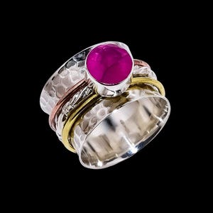 Può includere: Un anello spinner argentato con una texture martellata. L'anello presenta una grande gemma rotonda color magenta al centro. Ha fasce di metallo dorato e ramato su sfondo nero.