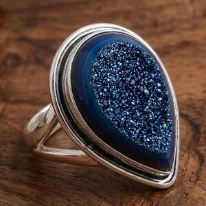 May include: A silver ring featuring a large, teardrop-shaped blue druzy stone. The stone has a sparkling, crystalline texture and is set in a polished silver band. The ring is displayed on a wooden surface.