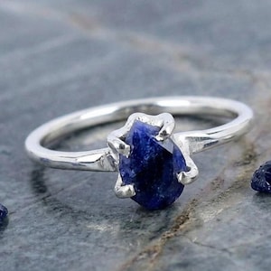 May include: A silver ring featuring a single, rough-cut, pear-shaped dark blue sapphire gemstone held in place by four metal prongs. The band is smooth and polished, showcasing a metallic luster that contrasts with the deep blue of the stone.
