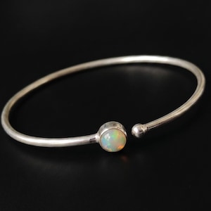 Op de afbeelding: Een zilverkleurige open armband met een ronde, iriserende opaal edelsteen. De armband heeft een eenvoudig, elegant ontwerp met een klein kogeldetail aan het open uiteinde. De opaal toont een mix van kleuren, waaronder wit, geel en groen.