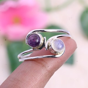 Op de afbeelding: Een zilveren ring met twee edelstenen. De ring heeft een gedraaid ontwerp en heeft een paarse amethist en een witte maansteen.