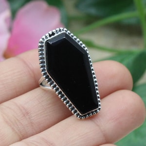 May include: A silver ring with a black coffin-shaped gemstone. The ring has a detailed, textured border.