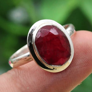 May include: A silver ring with a large, oval-shaped ruby gemstone. The ruby is a deep red color and has a faceted cut.