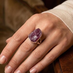 Peut inclure: Une bague avec une grosse pierre brute de couleur violette, sertie d'argent. La pierre a une surface rugueuse et textur&eacute;e, de forme irr&eacute;guli&egrave;re. La bague est port&eacute;e sur un doigt, mettant en valeur la taille et la couleur de la pierre.