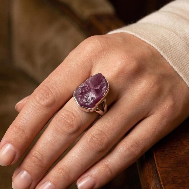 Peut inclure: Une bague avec une grosse pierre brute de couleur violette, sertie d'argent. La pierre a une surface rugueuse et textur&eacute;e, de forme irr&eacute;guli&egrave;re. La bague est port&eacute;e sur un doigt, mettant en valeur la taille et la couleur de la pierre.