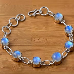 May include: A silver bracelet featuring round, iridescent moonstone cabochons. The stones are set in individual bezels and connected by a chain of interlocking links. The bracelet has a lobster clasp closure.