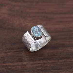 May include: A wide silver ring featuring a central oval-shaped light blue gemstone set in a smooth bezel. The band has a textured, hammered finish on the outer surface, creating a contrast with the polished edges and the clear blue stone.