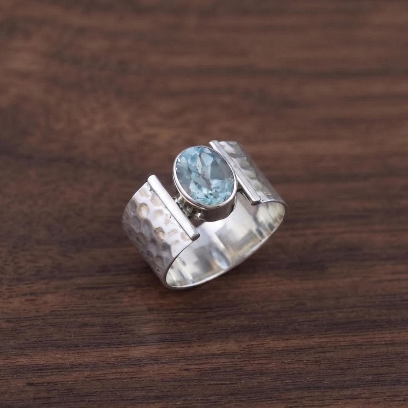 May include: A wide silver ring featuring a central oval-shaped light blue gemstone set in a smooth bezel. The band has a textured, hammered finish on the outer surface, creating a contrast with the polished edges and the clear blue stone.