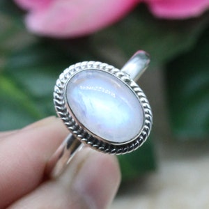 Puede incluir: Un anillo de plata con un cabujón ovalado de piedra lunar blanca engastado en un bisel de cuerda. El anillo está en un dedo.
