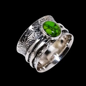 Peut inclure: Une bague en argent ornée d'une pierre ovale vert vif. La bande de la bague est ornée d'un motif floral complexe et présente une surface polie. La pierre est sertie dans un sertissage à lunette, sur fond noir.