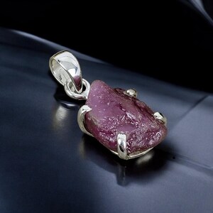 May include: A silver pendant featuring a raw pink ruby gemstone set in a silver bezel. The ruby has a rough, natural texture and is a deep pink colour.