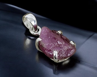 Puede incluir: Un colgante de plata con una piedra preciosa de rubí rosa en bruto engastada en un engaste de plata. El rubí tiene una textura rugosa y natural y es de color rosa intenso.