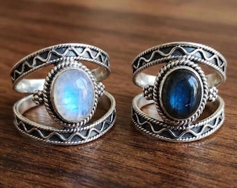 May include: Two silver rings featuring a double-band design with a zigzag pattern. The left ring holds an oval iridescent moonstone, while the right ring features an oval dark blue labradorite stone, both set in a beaded metal frame.